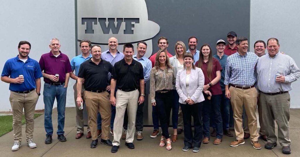 A group of employees posing outside their facility in front of a large TWF sign, smiling for a team photo.